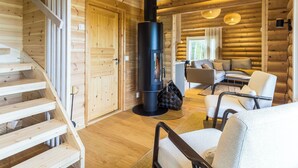 A log cabin interior with a central fireplace, wooden walls, a staircase, and a seating area.