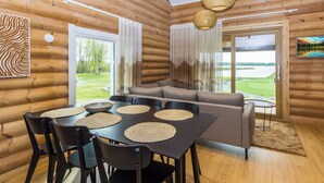 A log cabin interior with a dining area, sofa, and large windows overlooking a lake.