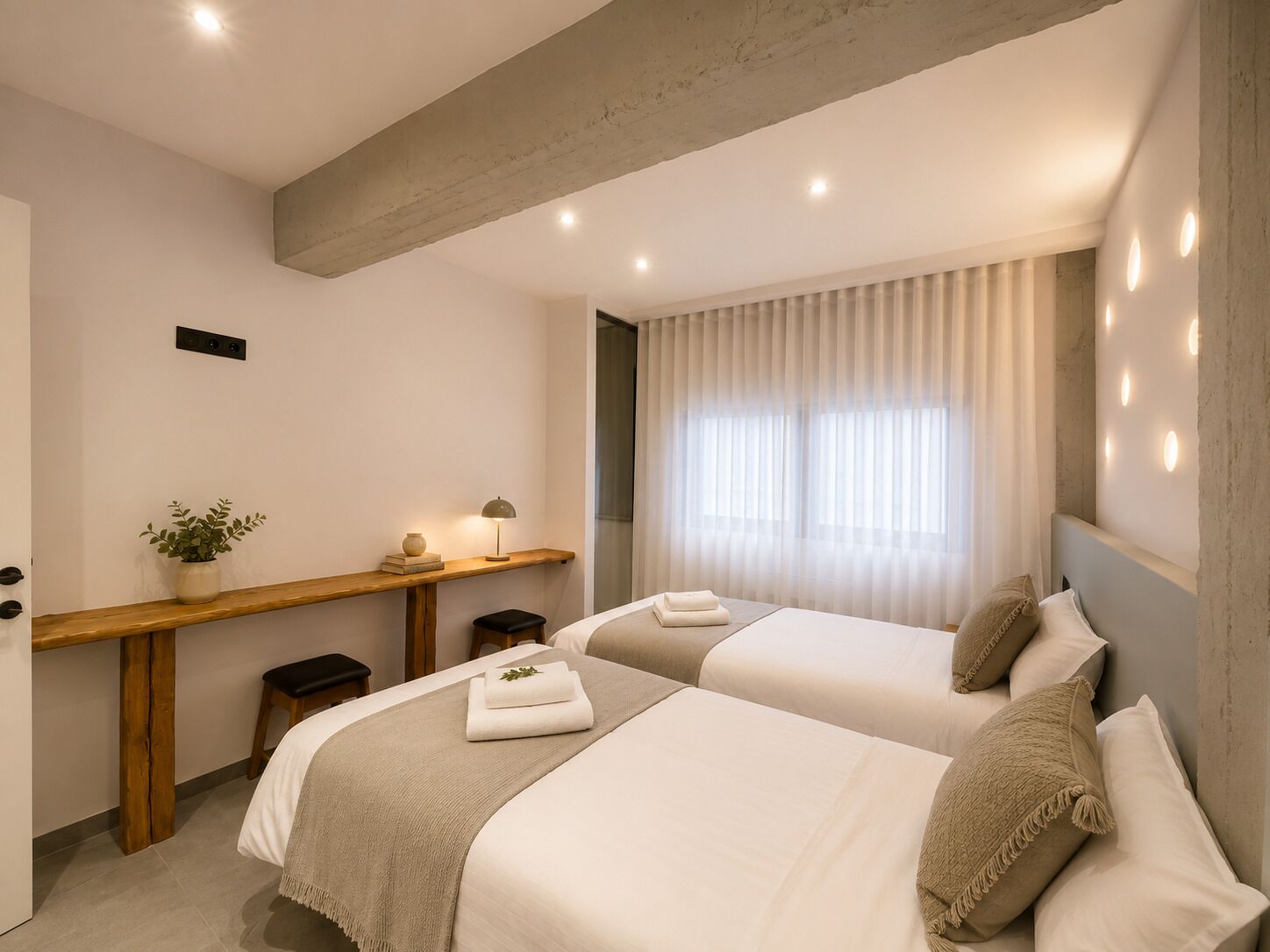Ein modernes Hotelzimmer mit zwei Betten, einem Schreibtisch mit Pflanze, einem kleinen Hocker und einem Fenster mit Vorhängen.