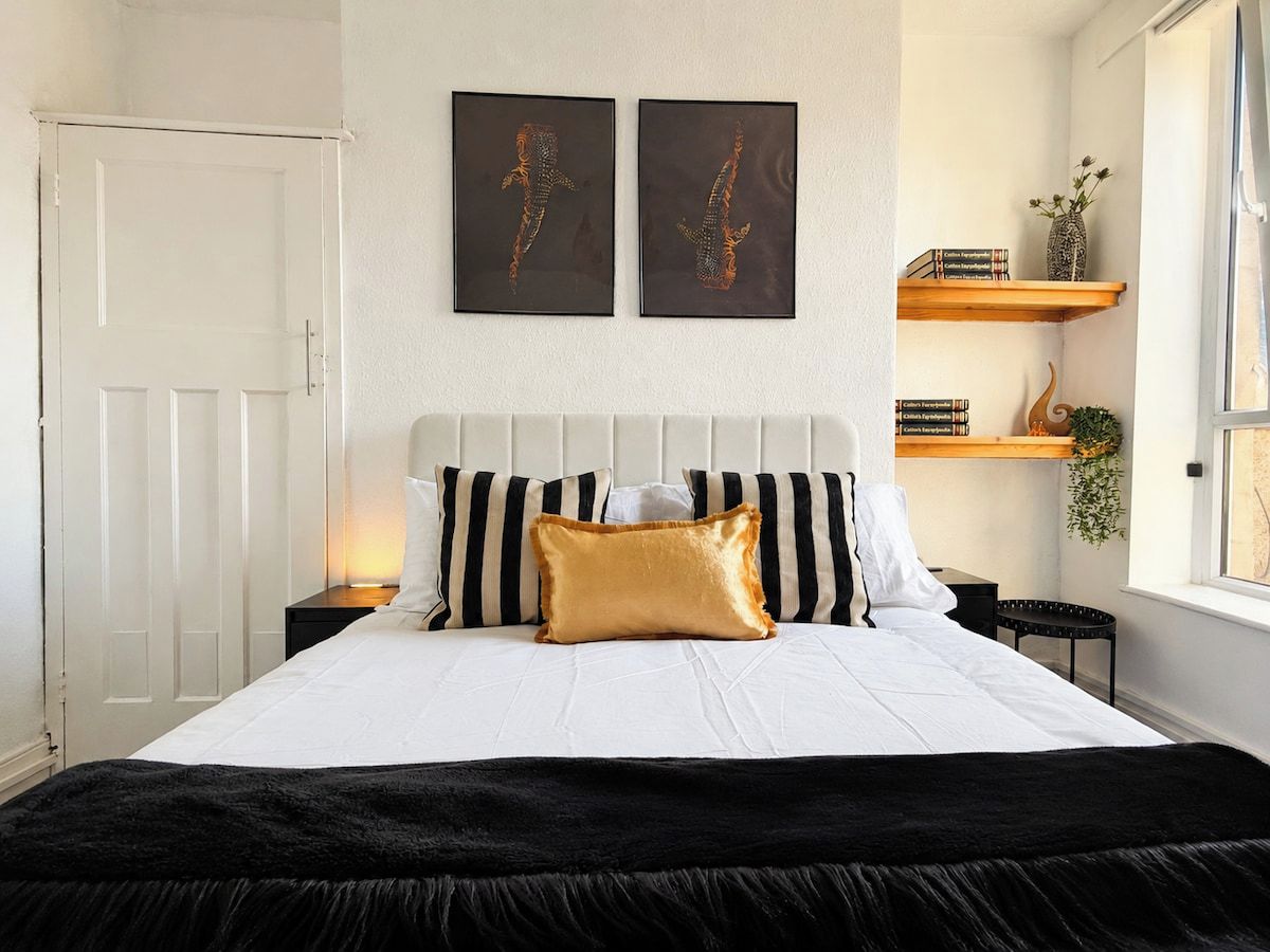 Ein Bett mit grau und weiß gestreifter Kopfteil, einem goldenen Akzentkissen und einer schwarzen Tagesdecke. Darüber an der Wand hängen zwei gerahmte Kunstwerke.