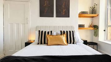 Ein Bett mit grau und weiß gestreifter Kopfteil, einem goldenen Akzentkissen und einer schwarzen Tagesdecke. Darüber an der Wand hängen zwei gerahmte Kunstwerke.