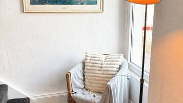 Een hoekkamer met een lijstwerk schilderij, een fauteuil met plaid, een vloerlamp en een raam.