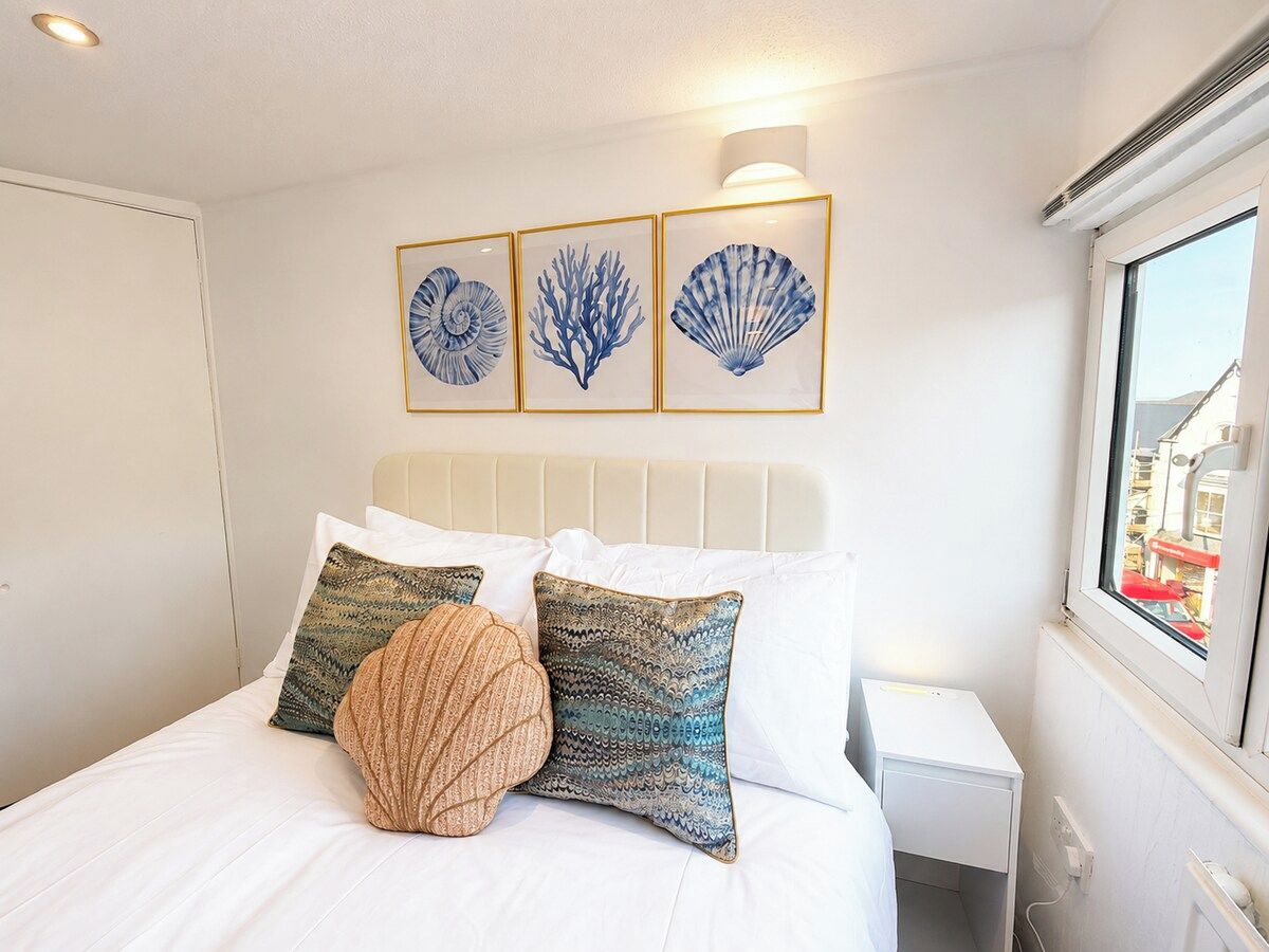 A neatly made bed with white bedding and decorative pillows, a headboard with a light fixture, and framed artwork on the wall.