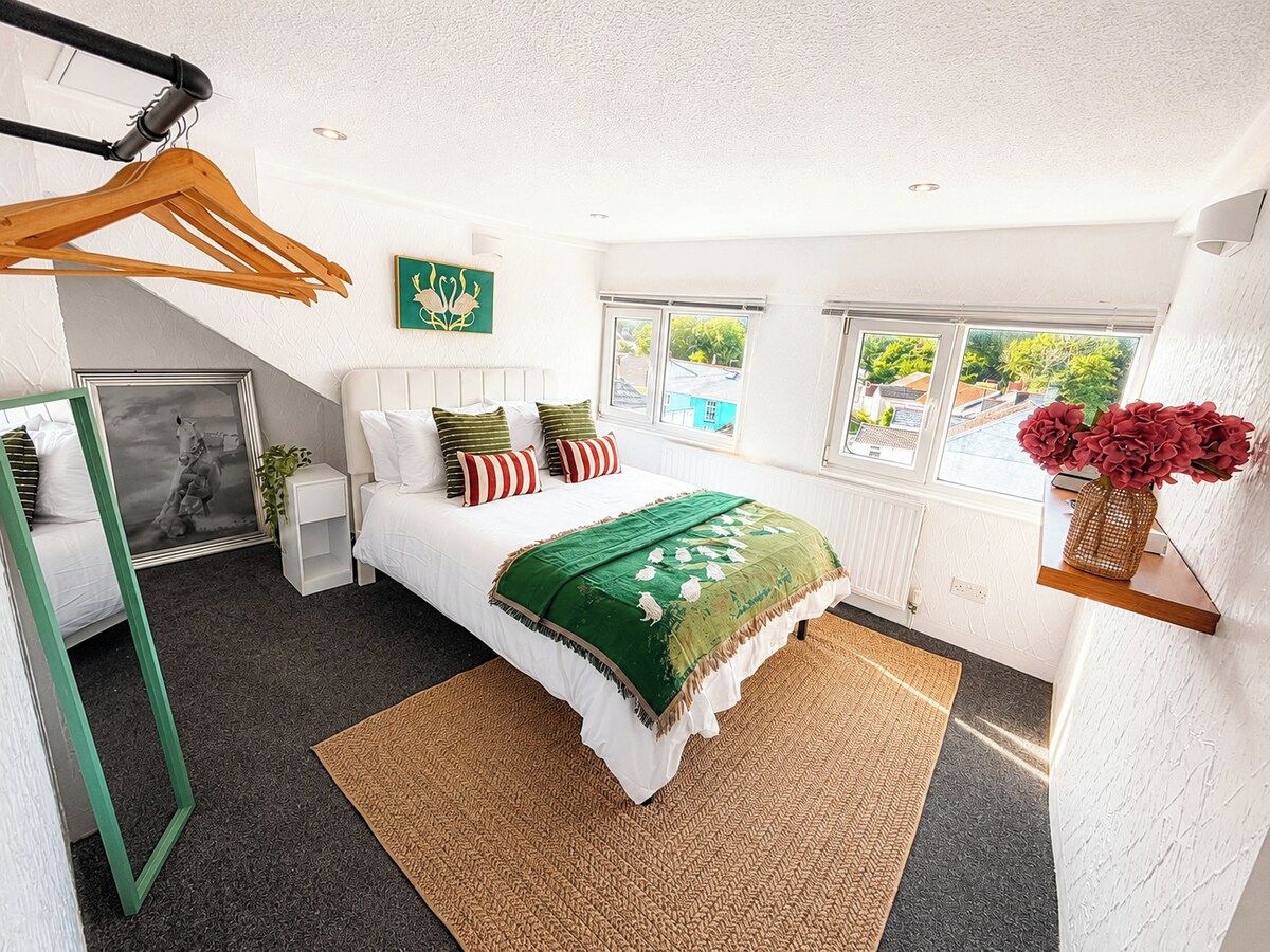A bedroom with a bed, bedside tables, a hanging coat rack, a window with a view, and a small shelf with flowers.