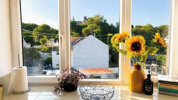 Ein Fenster mit Blick auf ein weißes Gebäude und Bäume, eine Vase mit Sonnenblumen, eine Packung Taschentücher, ein schwarzer Seifenspender und eine Metallschale auf der Fensterbank.