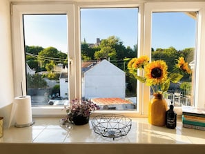A window with a view of a white building and trees, a vase with sunflowers, a tissue roll, a black soap dispenser, and a metal bowl on the windowsill.