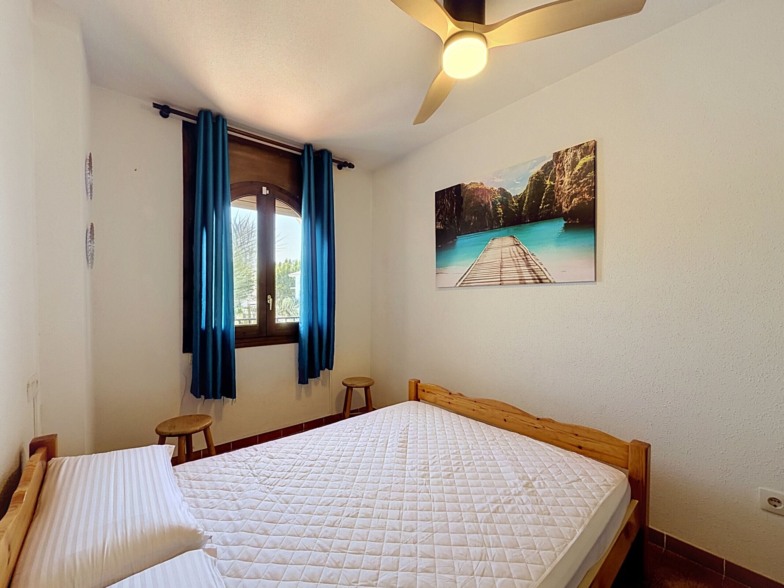 Une chambre à coucher avec un lit, une fenêtre avec des rideaux, un ventilateur de plafond et un tableau accroché au mur.