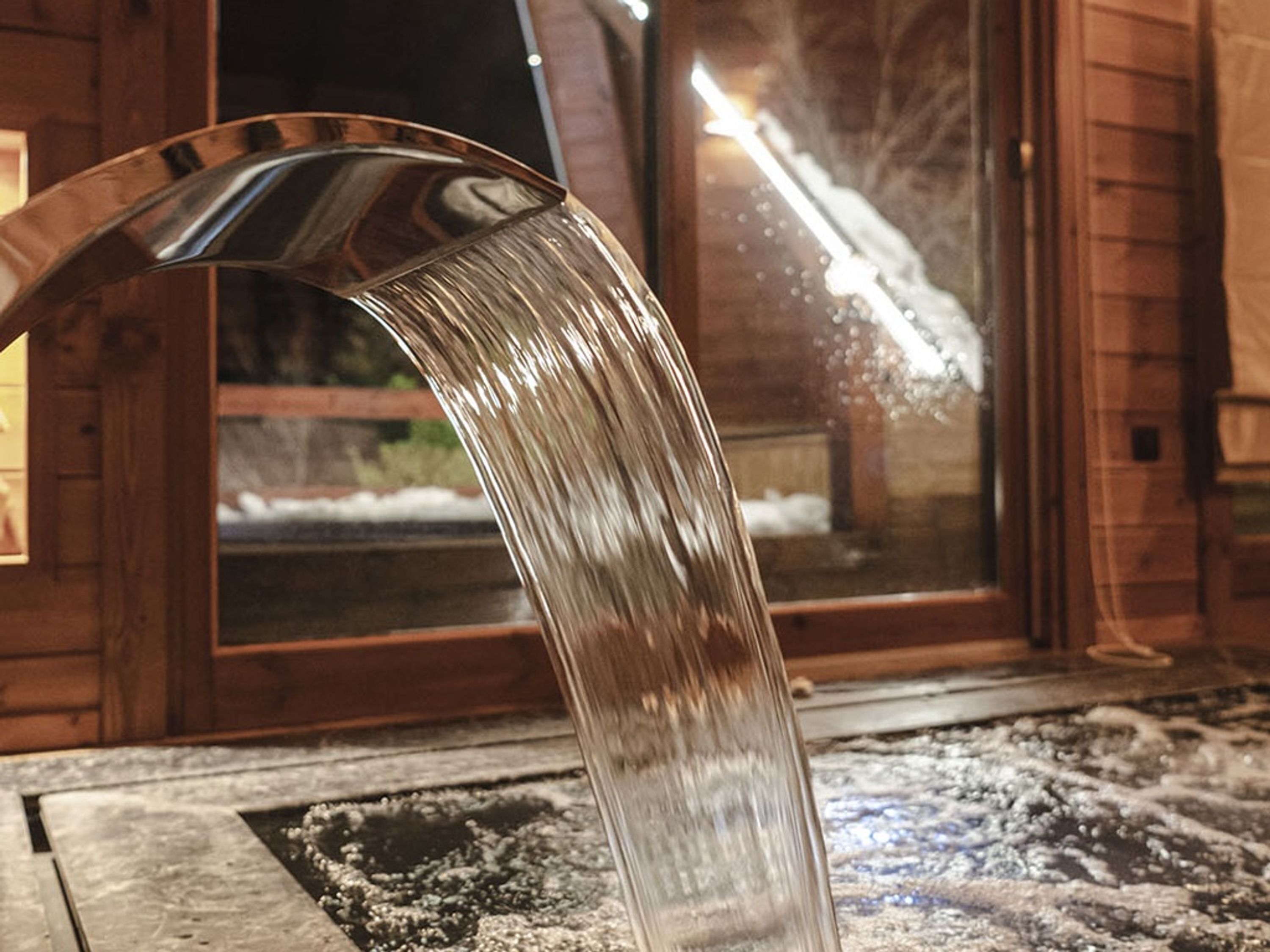 Une gouttière métallique d’où s’écoule de l’eau sur une zone enneigée, à l’extérieur d’un bâtiment en bois.