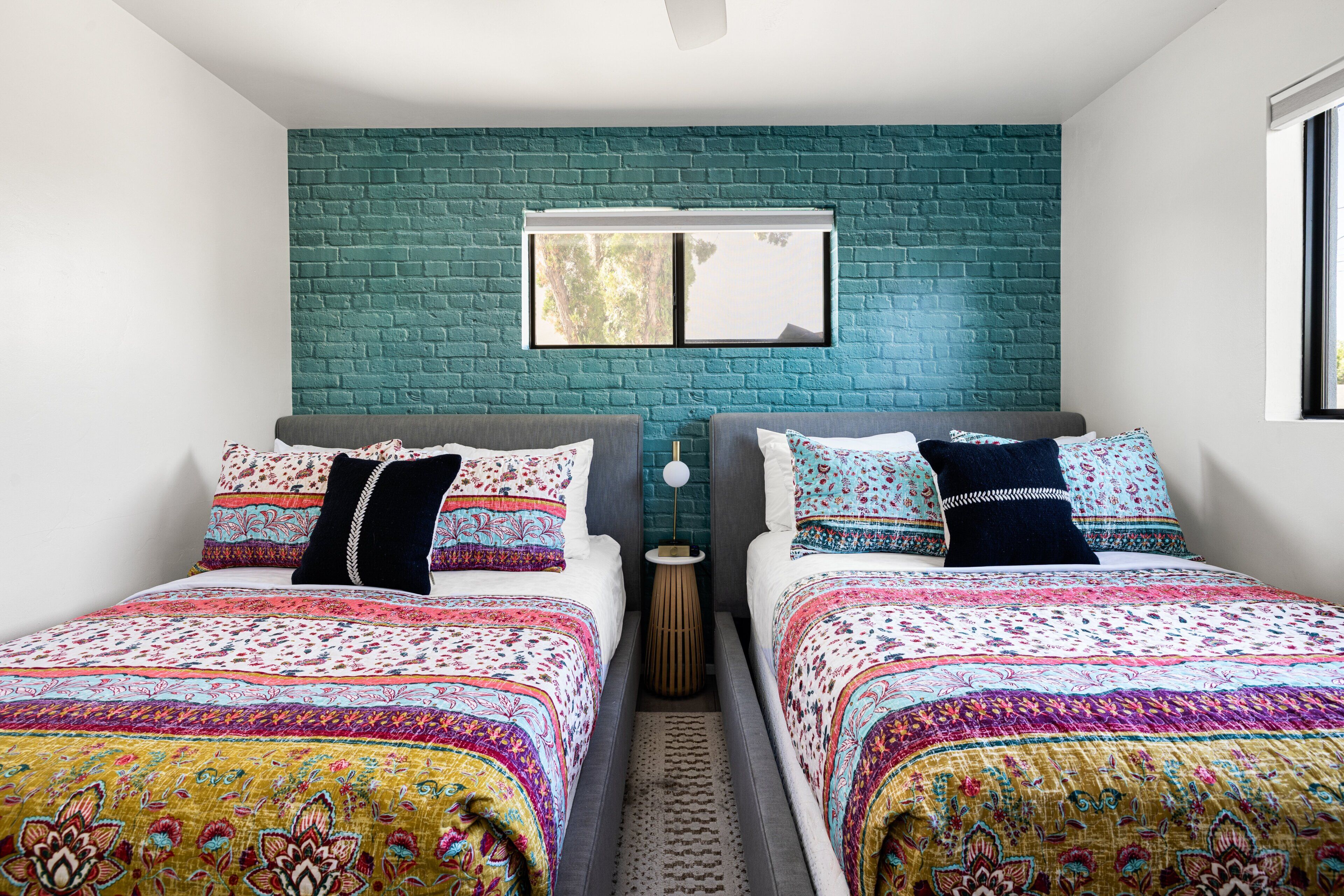A bedroom with two beds, a teal brick wall, a window, and a small bedside table.