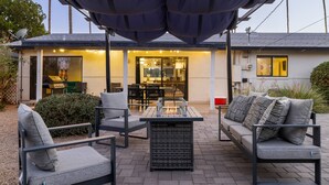 Eine Terrasse im Freien mit überdachter Sitzgruppe, einem Feuerbereich und Blick ins Hausinnere.