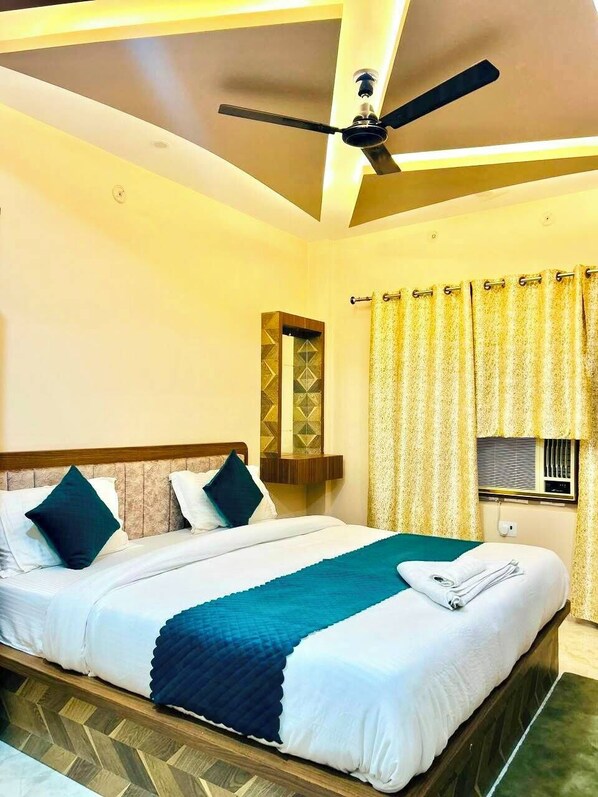 A hotel room with a bed, a ceiling fan, a window with curtains, and a wall-mounted air conditioning unit. - Hotel The Pushpraj (Varanasi)
