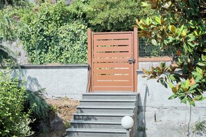 Unos escalones de concreto llevan a una verja marrón con listones horizontales, rodeada de follaje y una cerca.