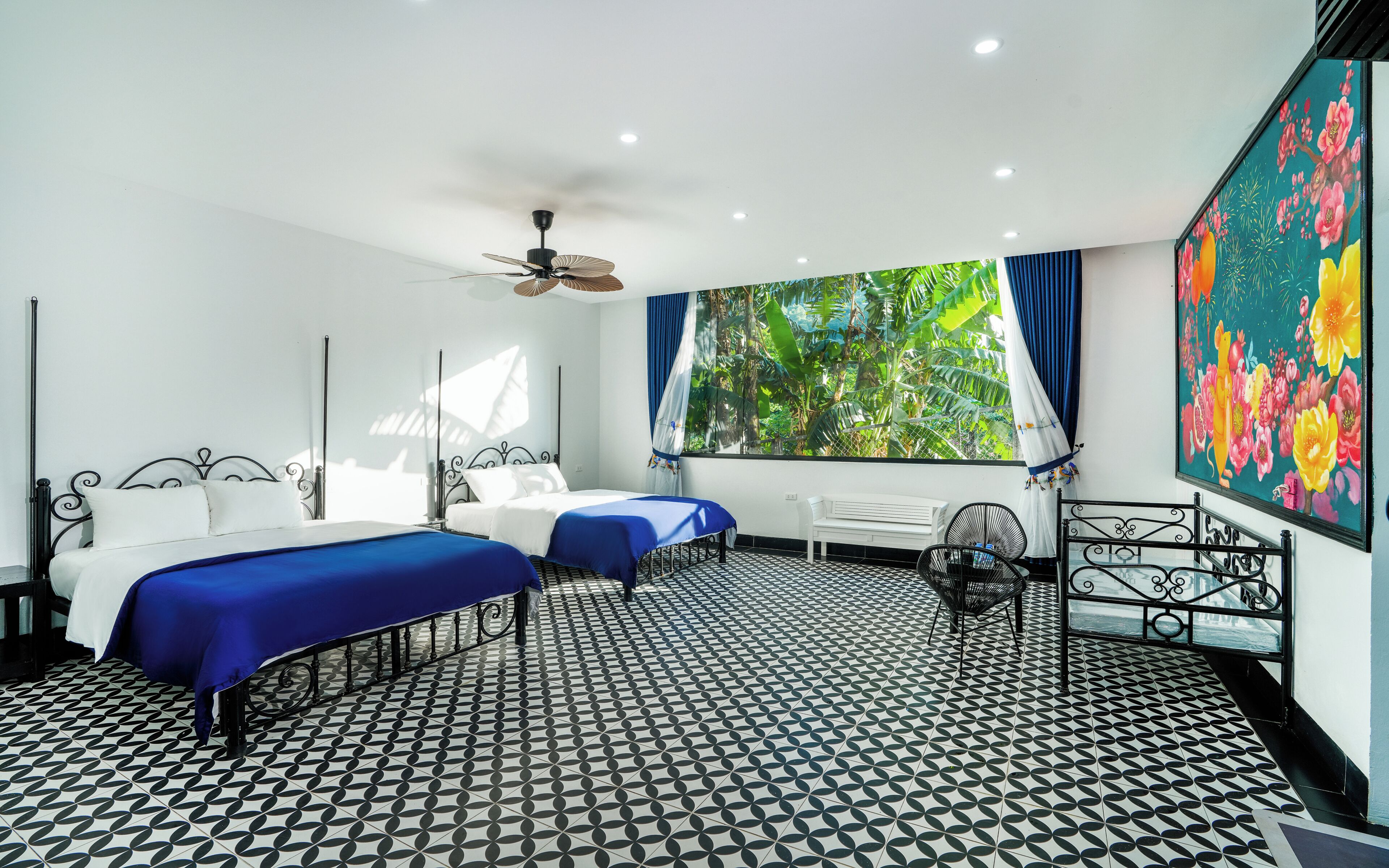 Une chambre d’hôtel avec deux lits, un ventilateur de plafond, une grande fenêtre avec des rideaux, un radiateur et un tableau accroché au mur.