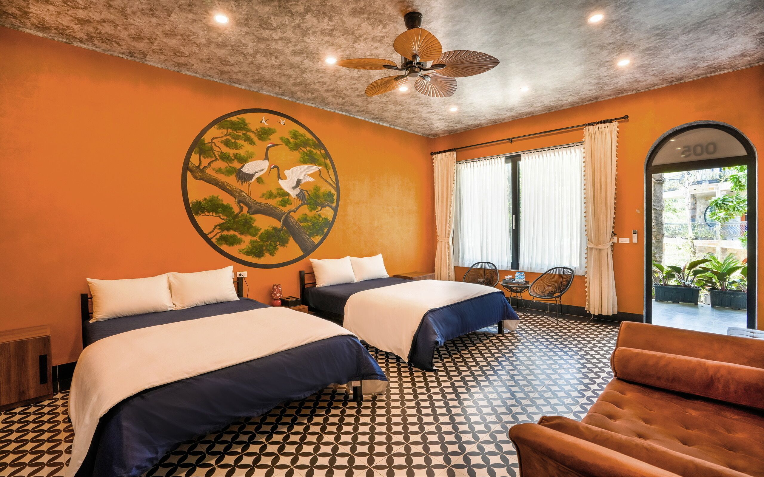 Habitación de hotel con dos camas, un ventilador de techo, un ventanal grande con cortinas y un suelo estampado.