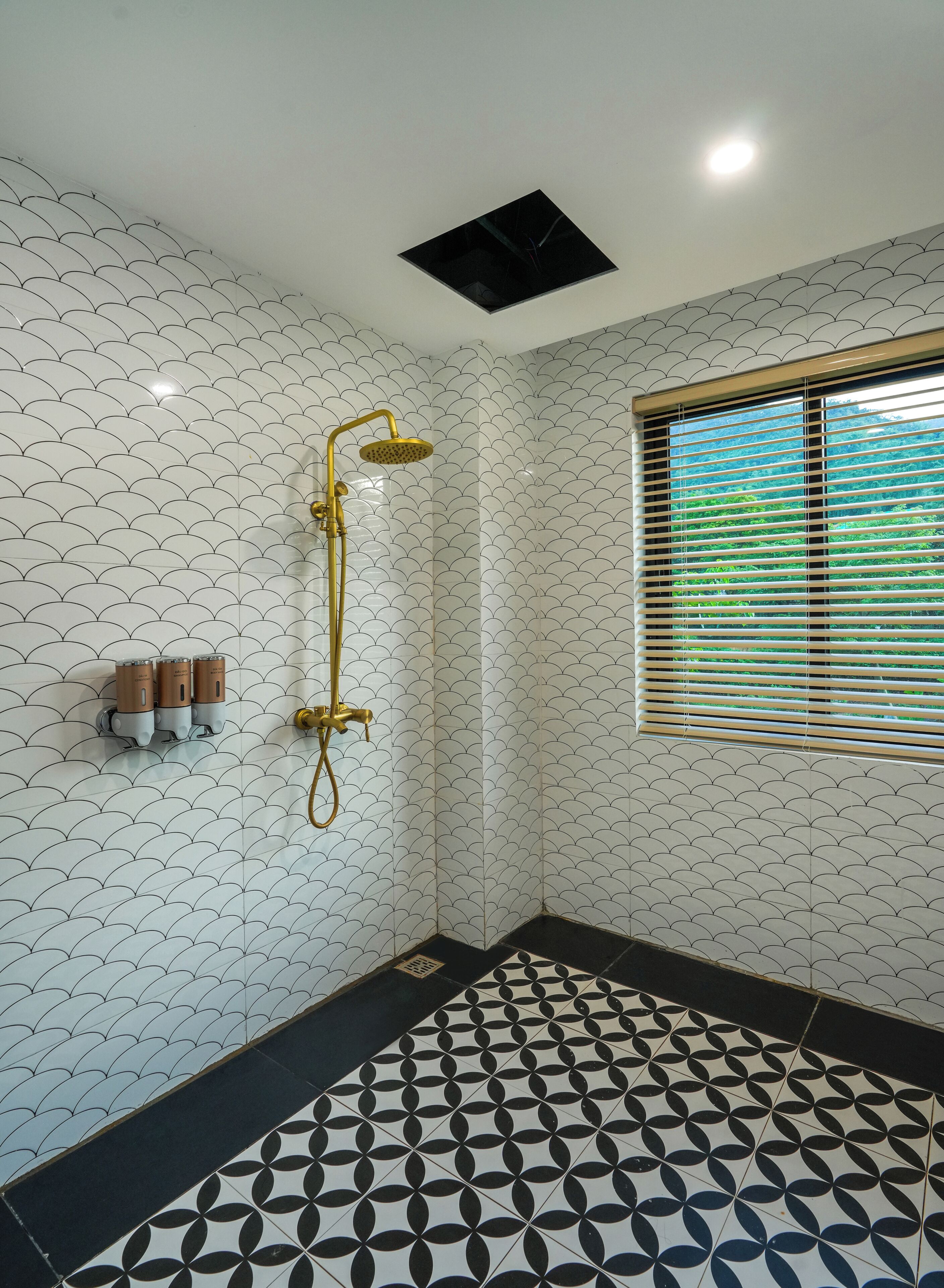 A bathroom with a patterned tile wall, a shower with a gold fixture, a window with blinds, and a black and white patterned floor.