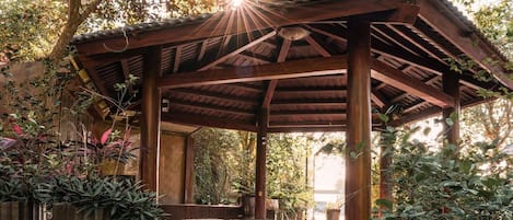 Un espacio al aire libre techado, con suelo de piedra, pilares de madera y plantas en macetas.
