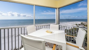 Un balcon avec une table et des chaises blanches, donnant sur une plage et l’océan.