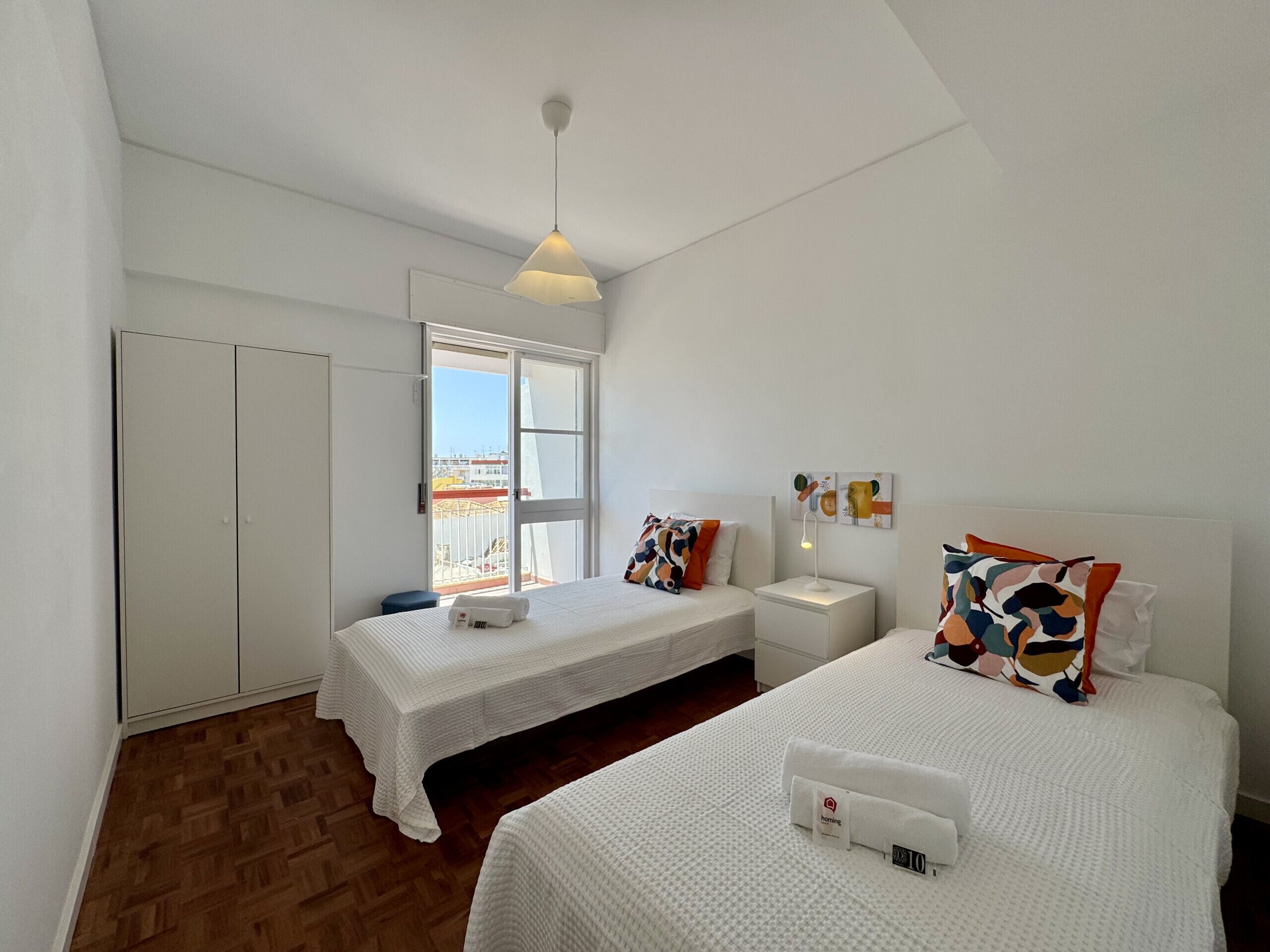 A hotel room with two beds, a wardrobe, a nightstand, a lamp, and a view of the outside.