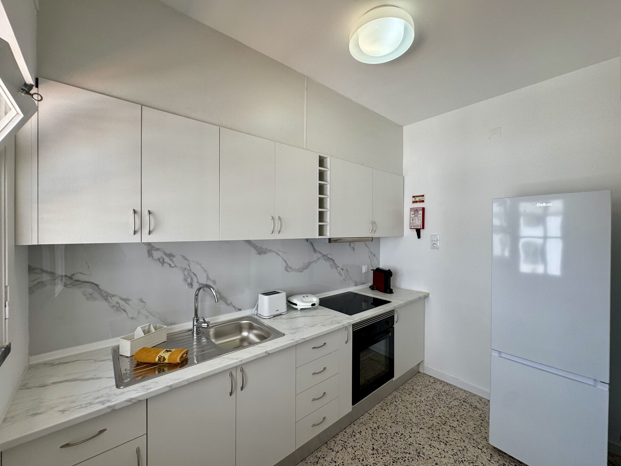 Una cucina con mobili bianchi, una parete di fondo in marmo, un lavello, un tostapane, un forno e un frigorifero.