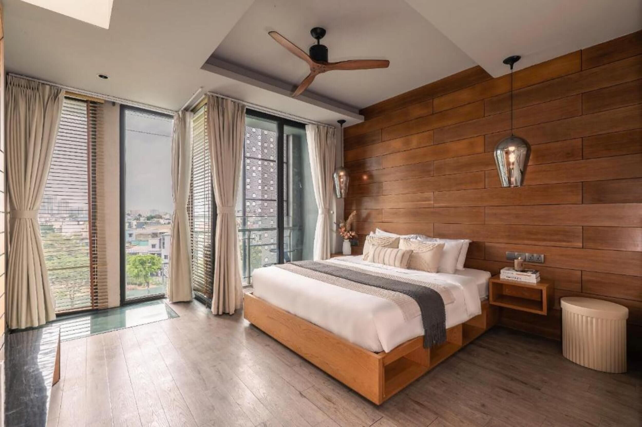Un dormitorio con cabecera de madera, cama con ropa blanca, mesita de noche, taburete redondo y ventilador de techo.