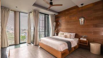 Un dormitorio con cabecera de madera, cama con ropa blanca, mesita de noche, taburete redondo y ventilador de techo.