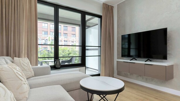 A modern living room with a sofa, a round table, a TV mounted on the wall, and large windows with curtains. - Beige and Blue Apartment | Garden | Parking (Gdańsk)