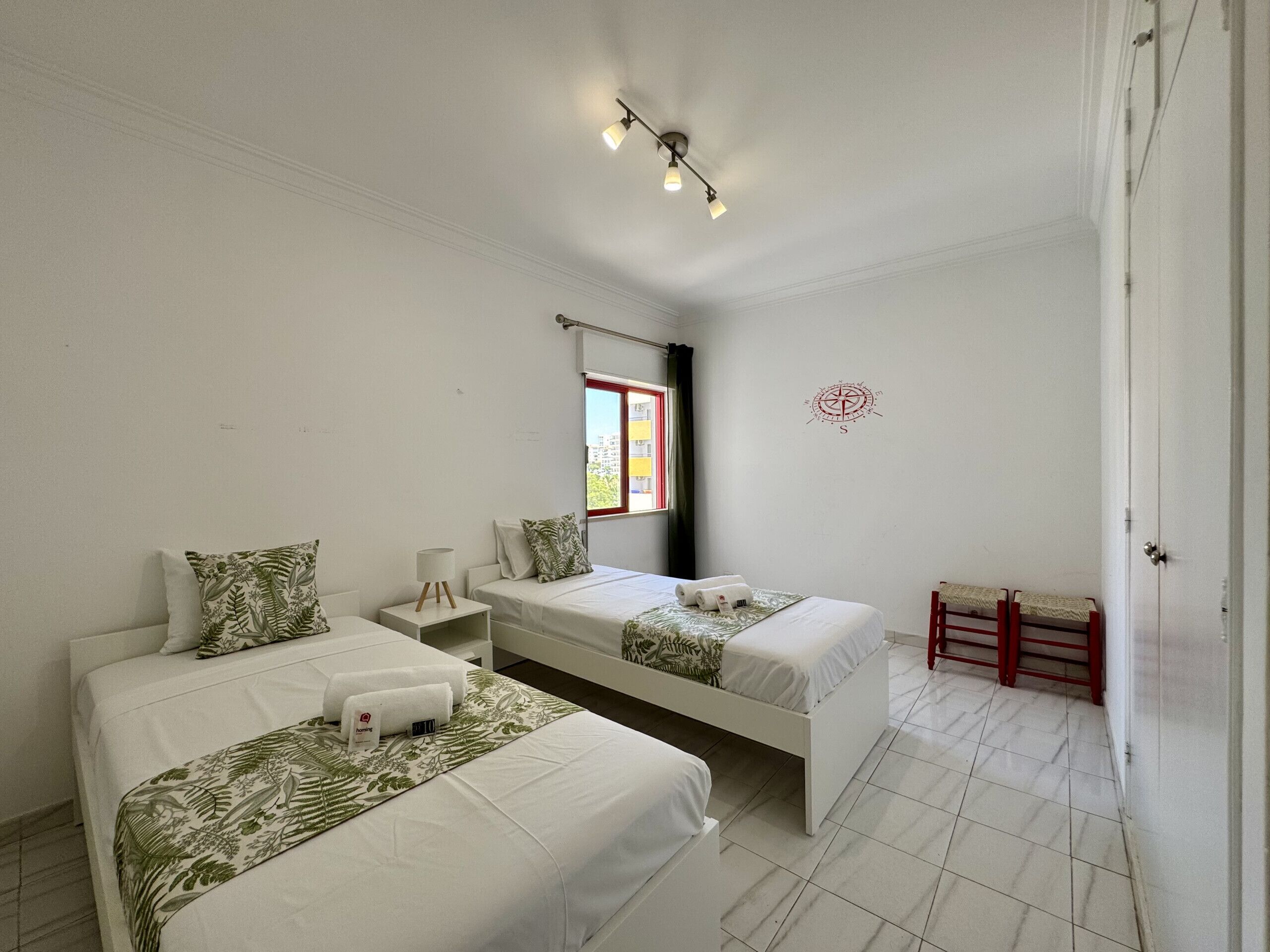 A hotel room with two beds, a small table, a nightstand, a window with a view, a red bench, and a compass wall decoration.