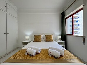 A hotel room with a bed, two bedside tables, a window with a view of buildings, and a patterned rug. - Quarteira Beach & Ocean View 3 by Homing (Loulé)