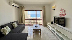 Ett vardagsrum med en soffa, ett soffbord, en tv och en balkong med utsikt över havet.