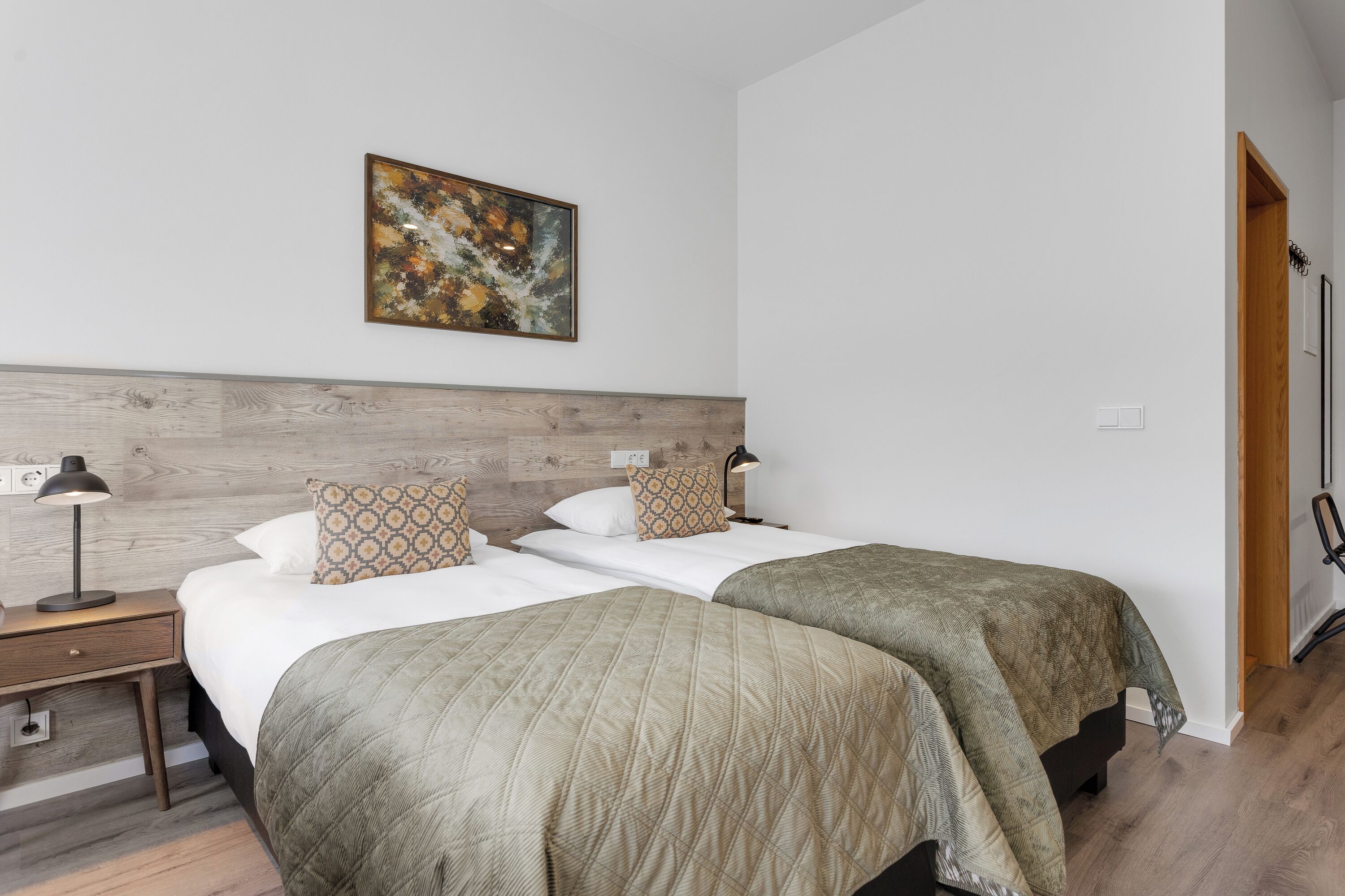 Una camera d'albergo con due letti, una testiera in legno, un comodino con una lampada e un quadro appeso al muro.