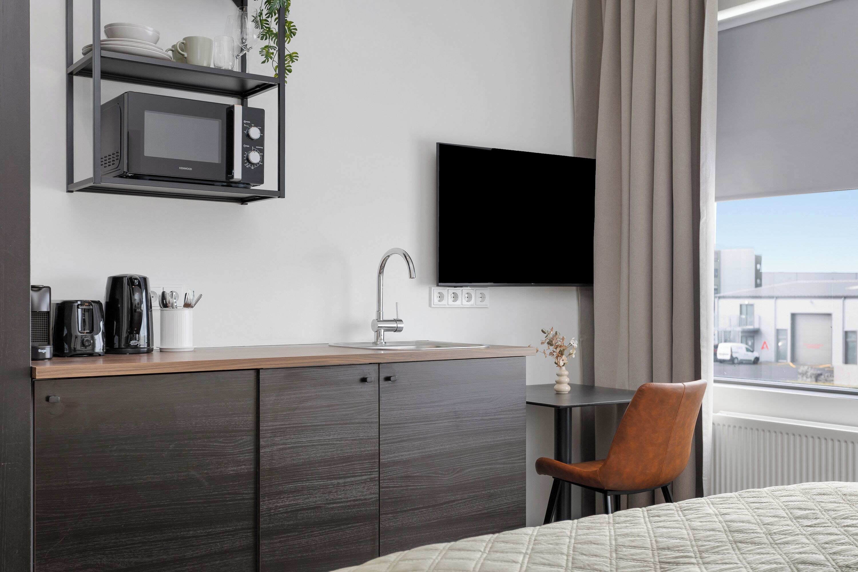 A modern hotel room with a kitchenette, a flat-screen TV, a window with a view of the city, and a bed with a quilted cover.