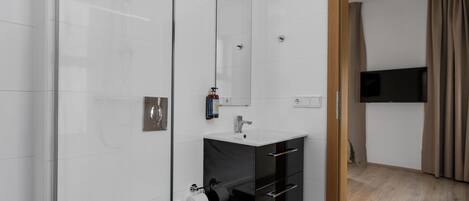 Un baño moderno con cabina de ducha de vidrio, inodoro blanco, tocador negro con espejo y televisor de pantalla plana montado en la pared.