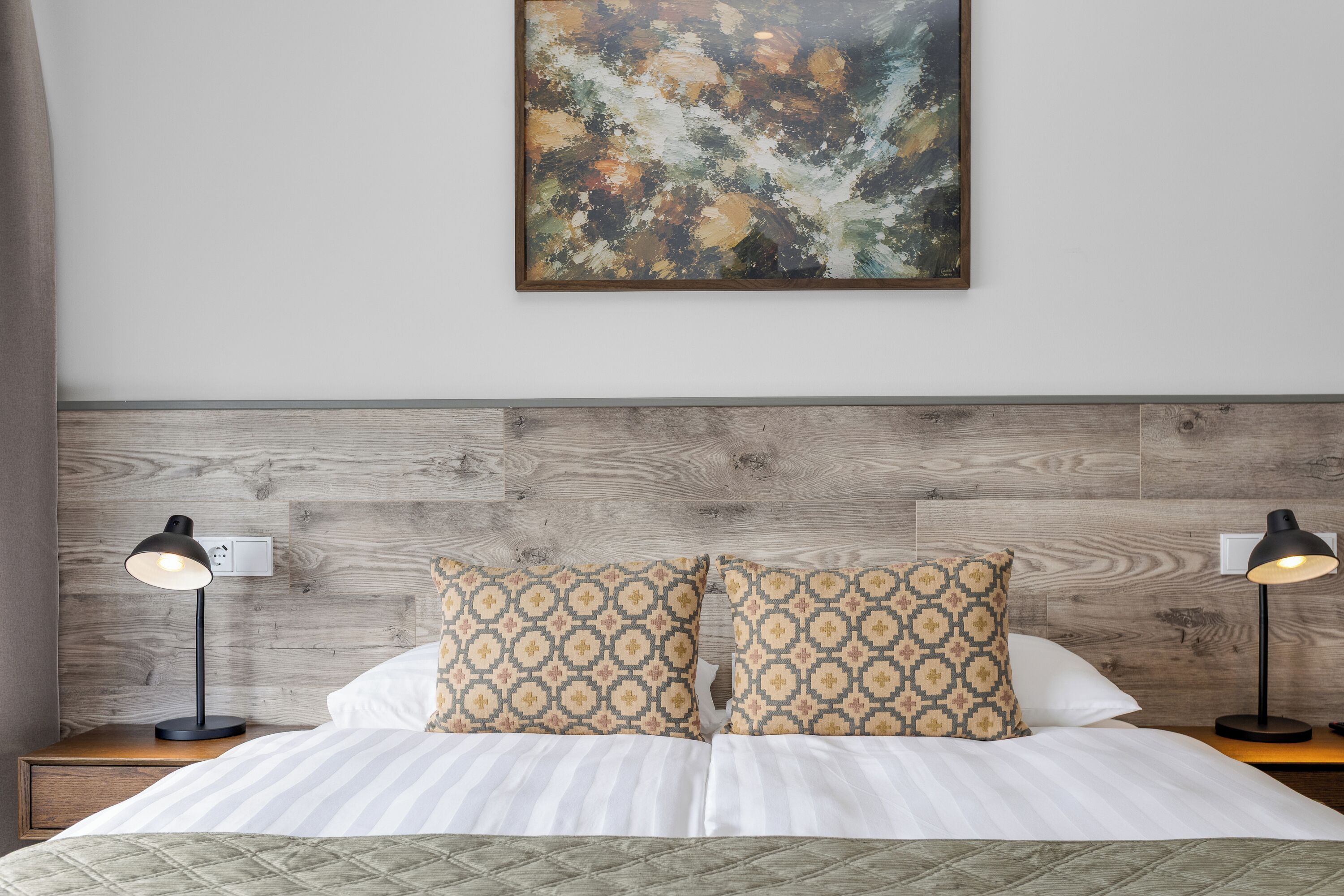 A hotel room with a wooden headboard, two bedside lamps, patterned pillows, a bed with white bedding, and a painting on the wall.