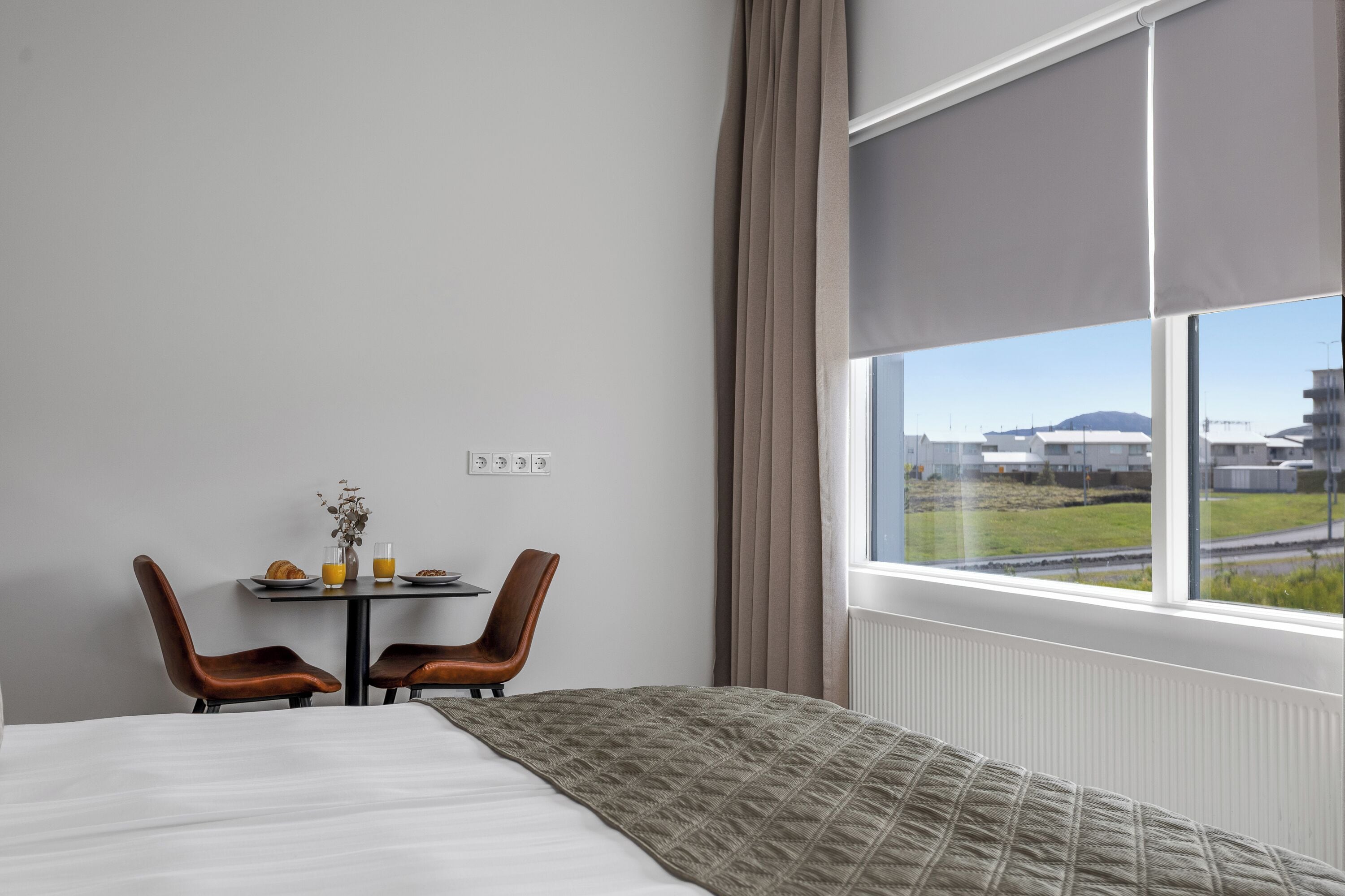 A hotel room with a bed, a table with breakfast items, two chairs, and a large window with a view of buildings.