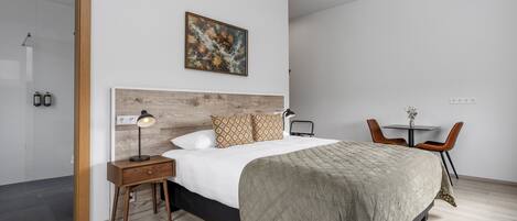 Un dormitorio moderno con cabecera de madera, una cama con manta verde acolchada, una mesita de noche con lámpara, una pequeña mesa con sillas y una zona de ducha.