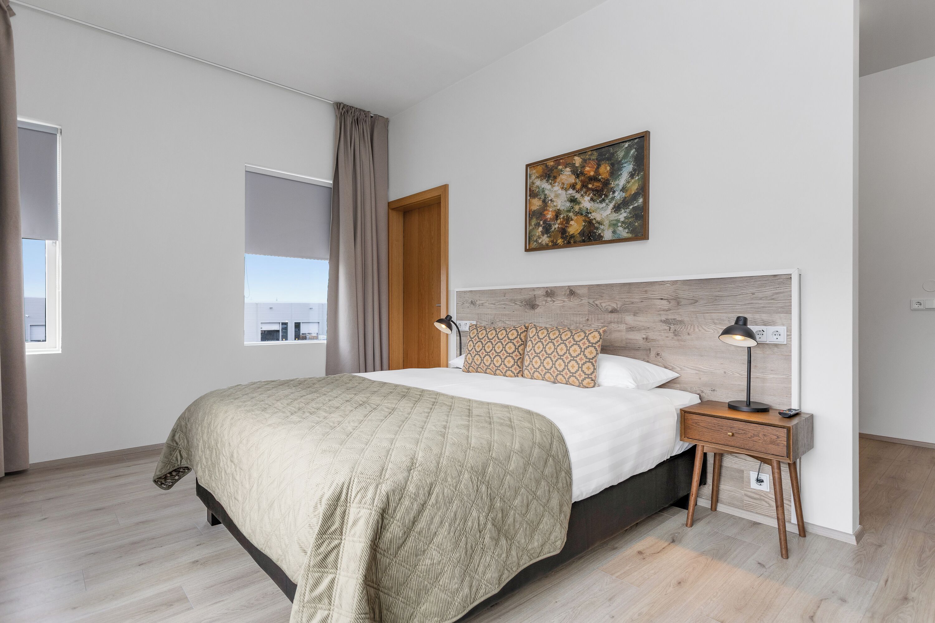 A modern bedroom with a large bed, a nightstand, a lamp, a painting, and a view through an open door.