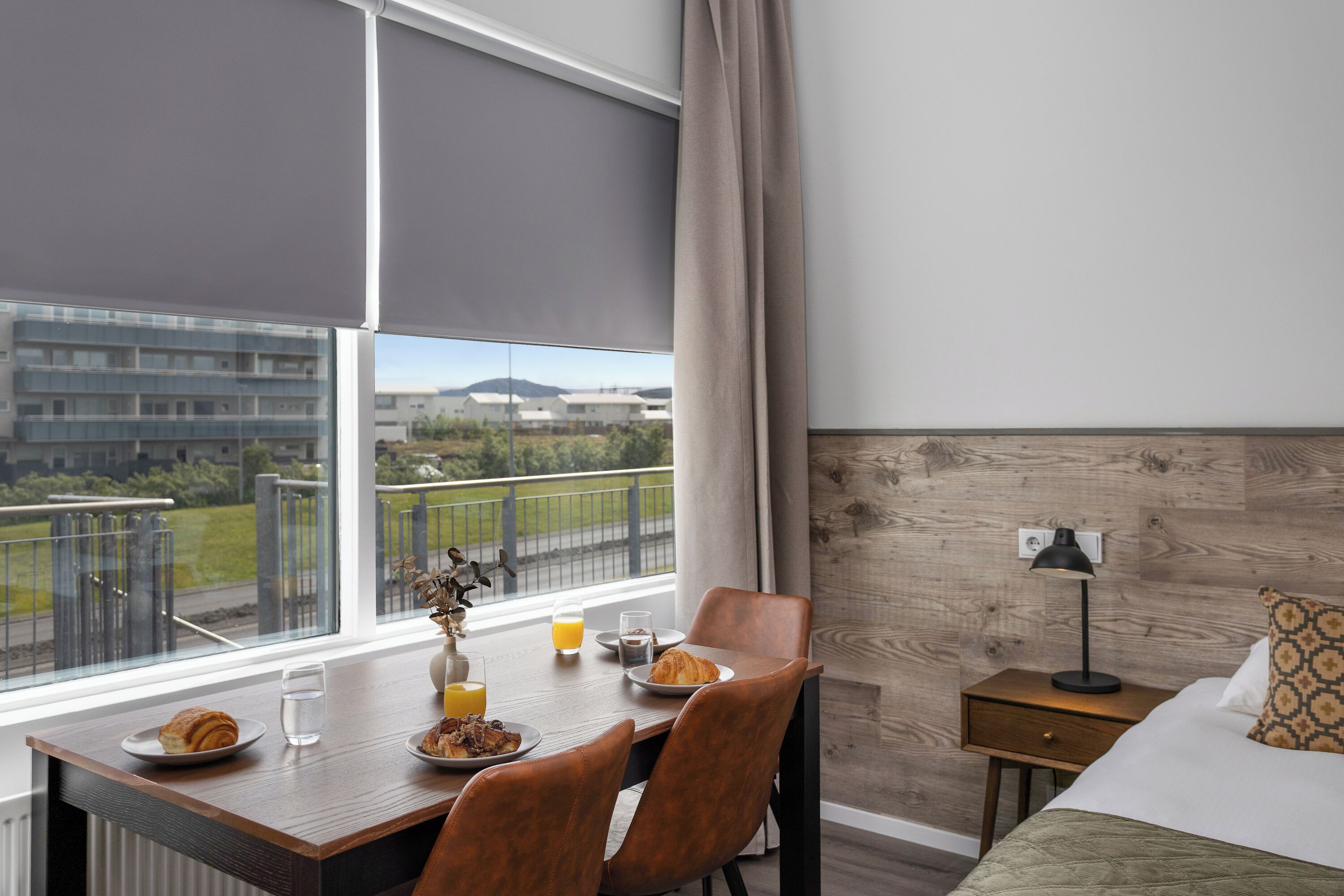 Habitación de hotel con una mesa de comedor lista para el desayuno, una cama con almohada estampada, una mesita de noche con lámpara y vistas a edificios y zonas verdes.