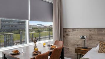 Habitación de hotel con una mesa de comedor lista para el desayuno, una cama con almohada estampada, una mesita de noche con lámpara y vistas a edificios y zonas verdes.