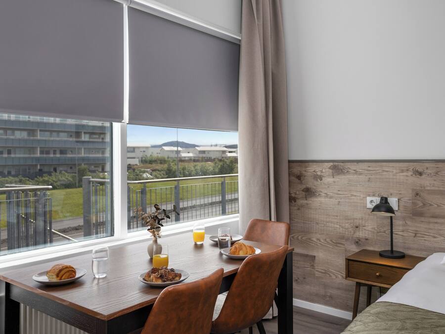 Habitación de hotel con una mesa de comedor lista para el desayuno, una cama con almohada estampada, una mesita de noche con lámpara y vistas a edificios y zonas verdes.