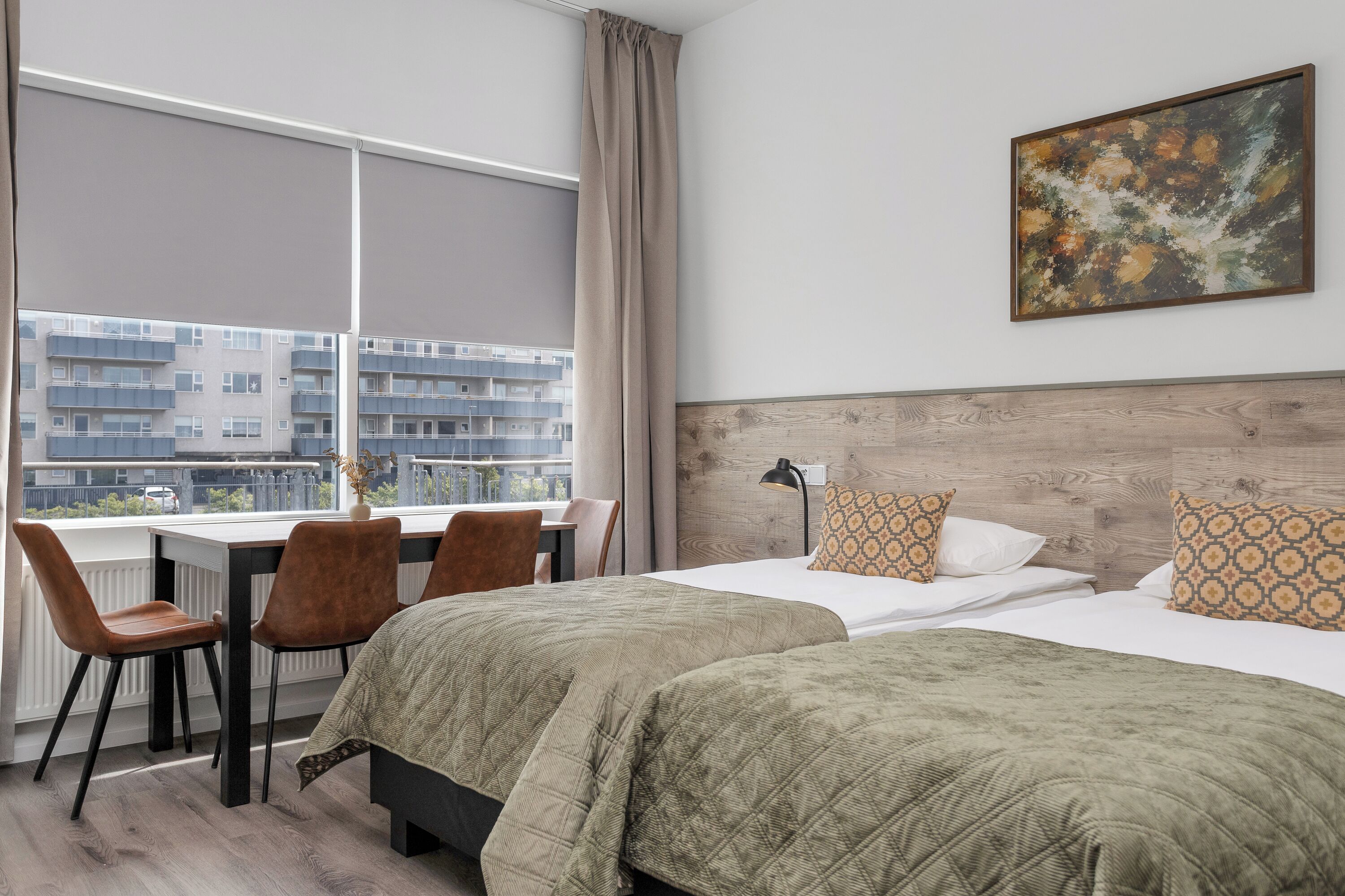 A hotel room with two beds, a desk, chairs, a window with curtains, a painting on the wall, and a view of buildings outside.