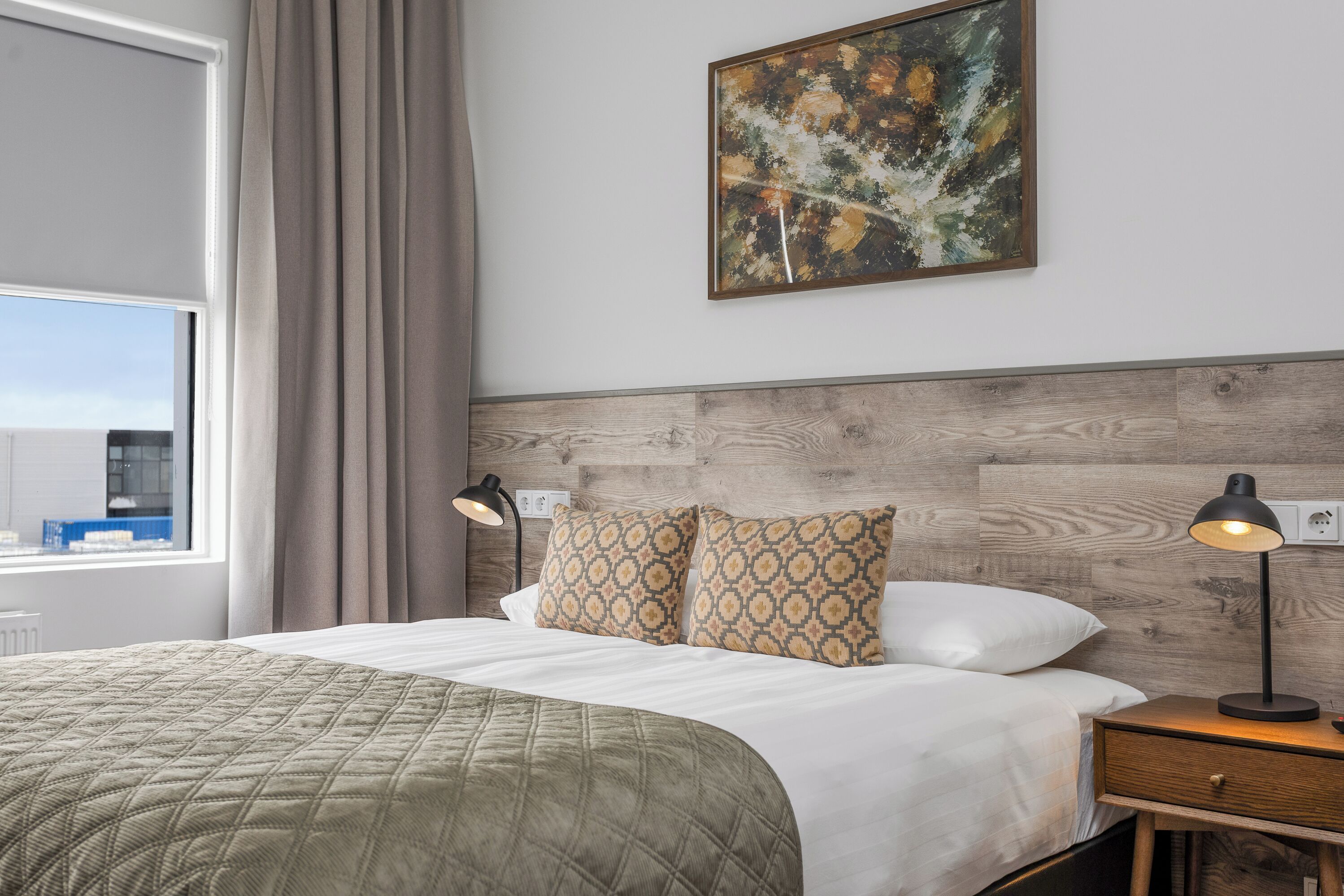 Hotellihuone, jossa on sänky, kaksi yöpöytälamppua, yöpöytä, laatikosto, ikkuna verhoilla ja taulu seinällä.