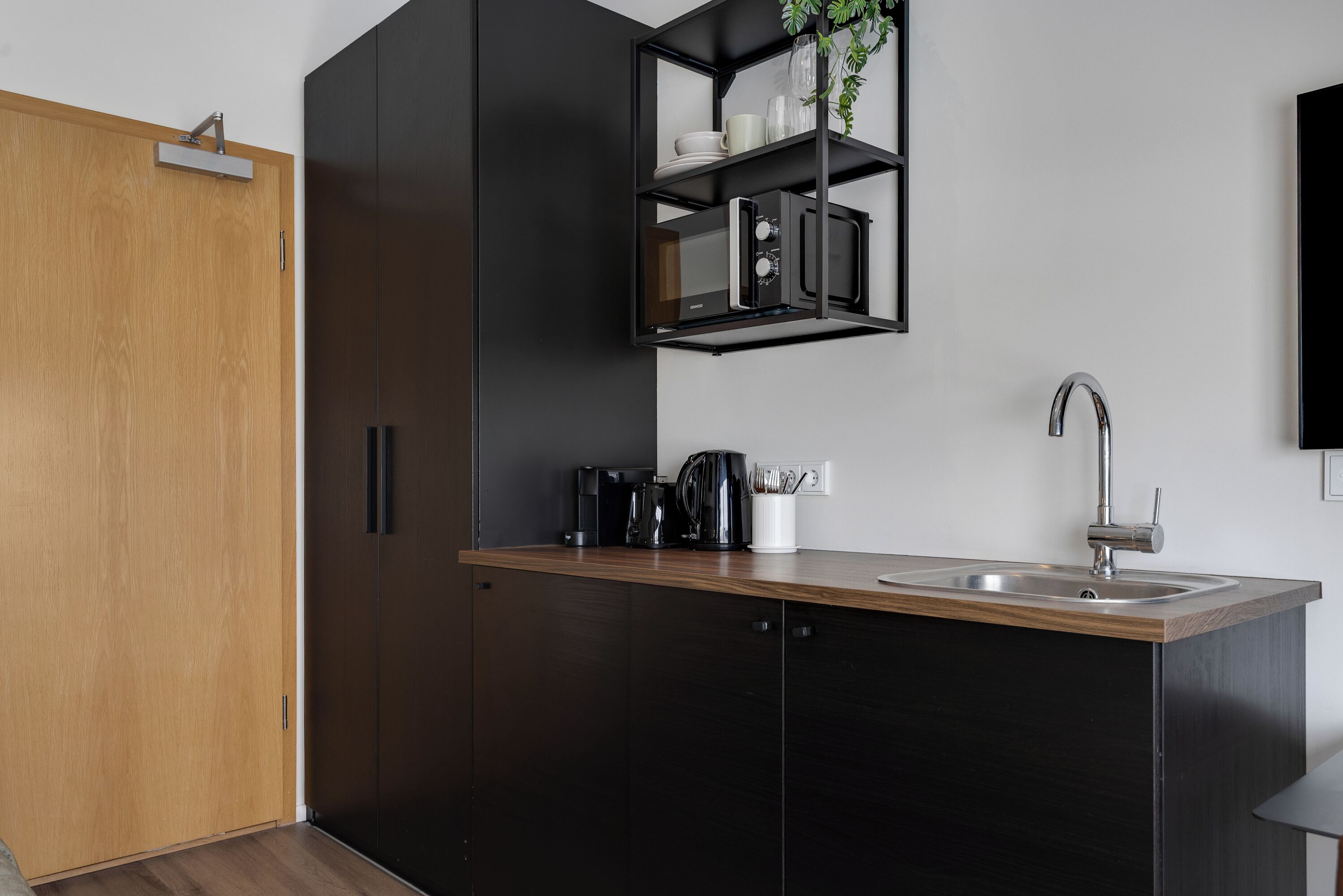 A modern kitchen with a wooden door, black cabinets, a stainless steel sink, and a black shelf with kitchen items.