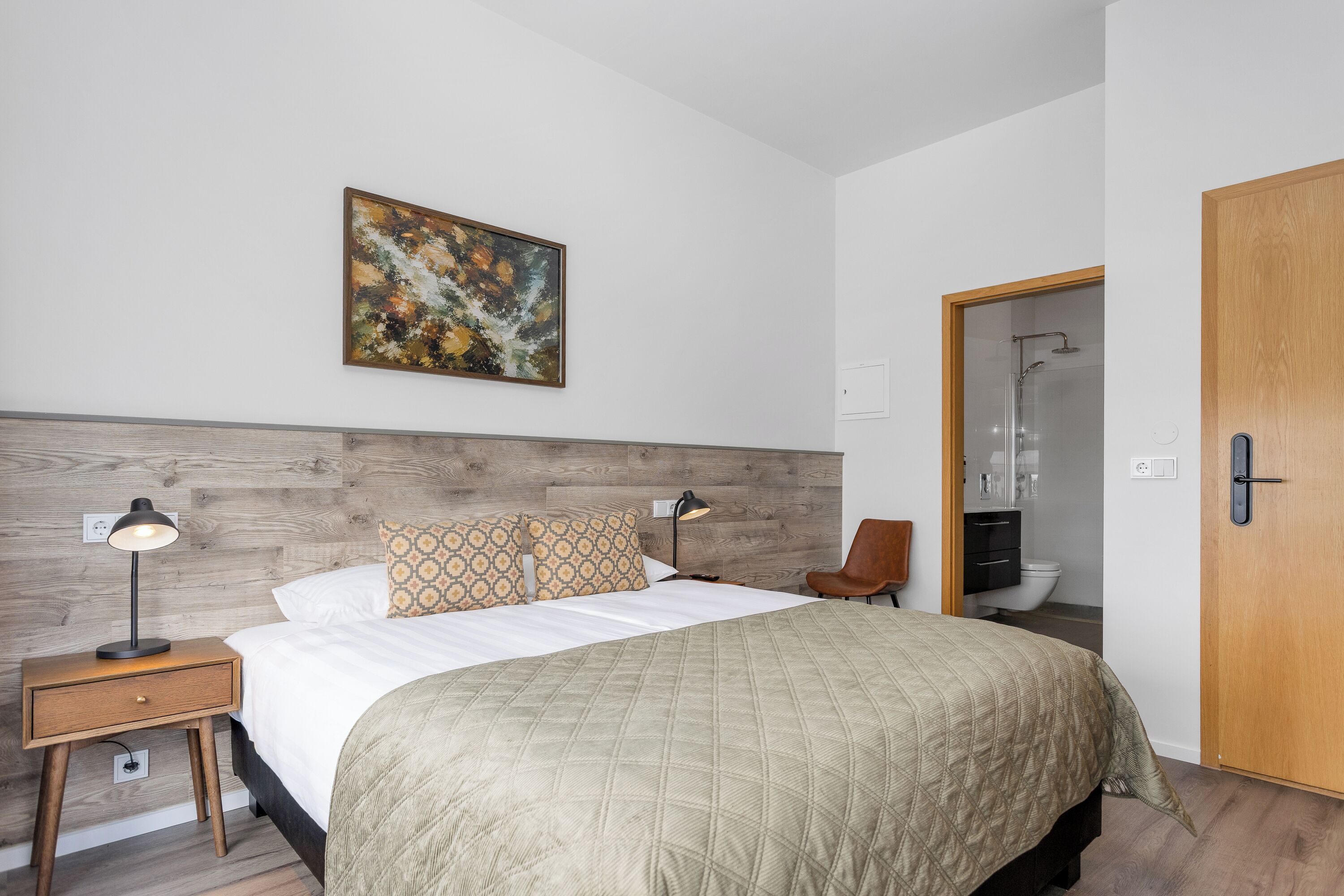 A bedroom with a bed, two bedside tables, a night lamp, a wooden headboard, a painting, a bathroom door, and a wooden floor.