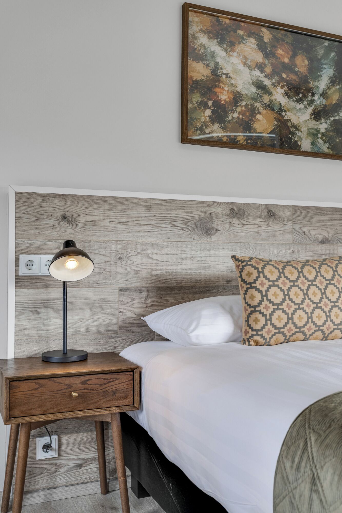 Un dormitorio con cabecera de madera, lámpara de noche, almohada estampada y un cuadro en la pared.