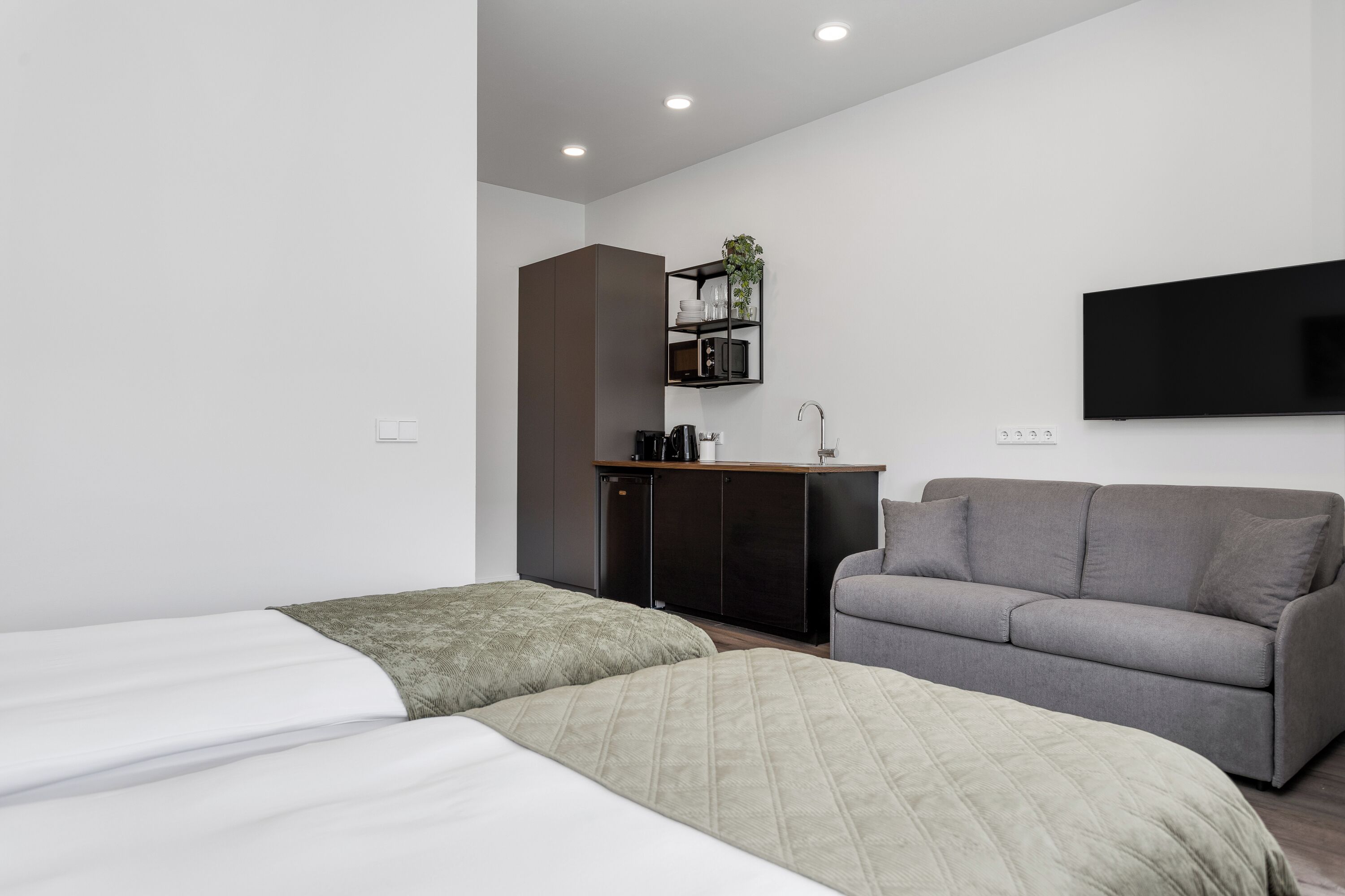 A modern hotel room with two beds, a gray sofa, a flat-screen TV, and a kitchenette.