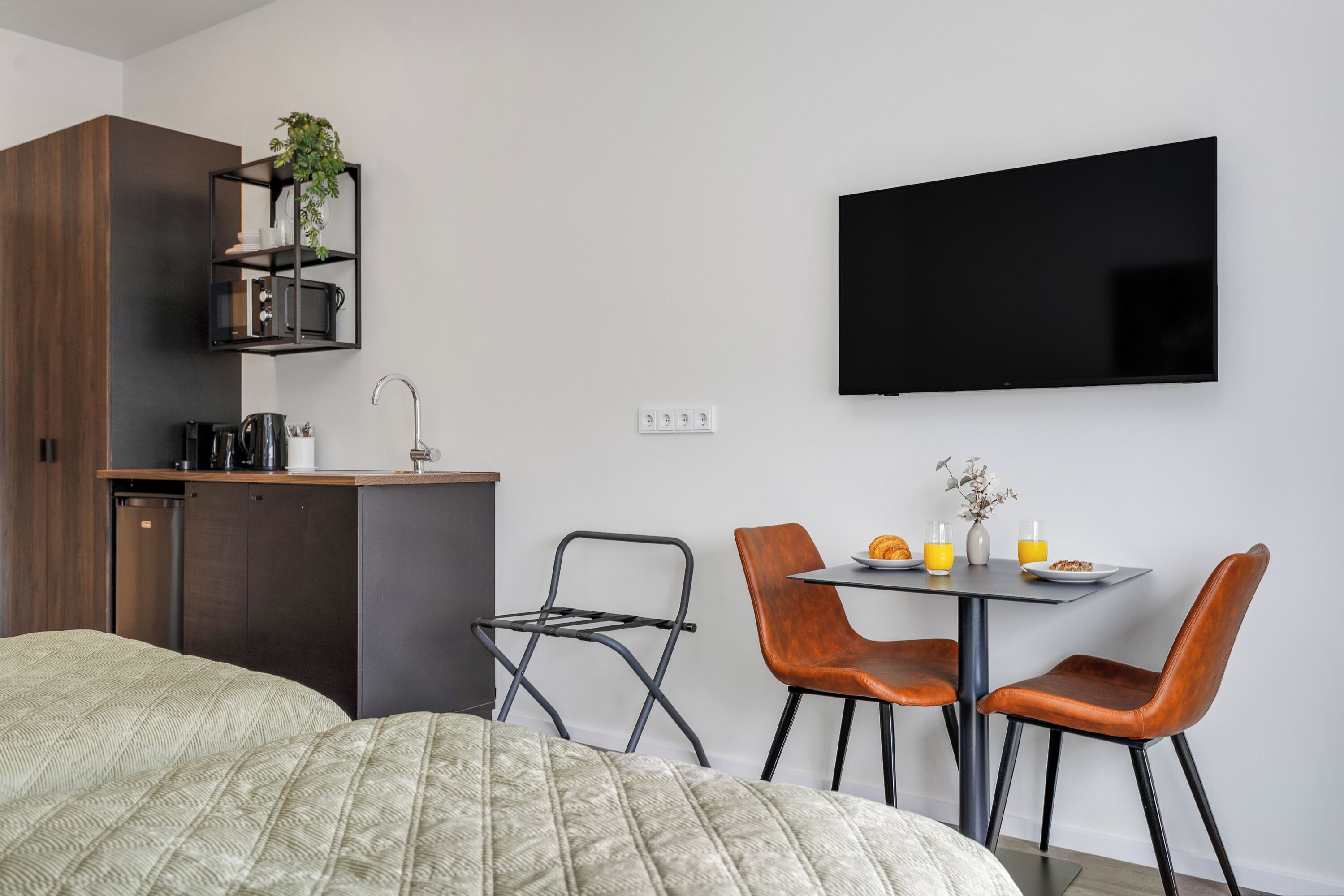 A modern hotel room with a bed, a kitchenette, a dining area with chairs, and a flat-screen TV.