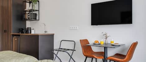 A modern hotel room with a bed, a kitchenette, a dining area with chairs, and a flat-screen TV.