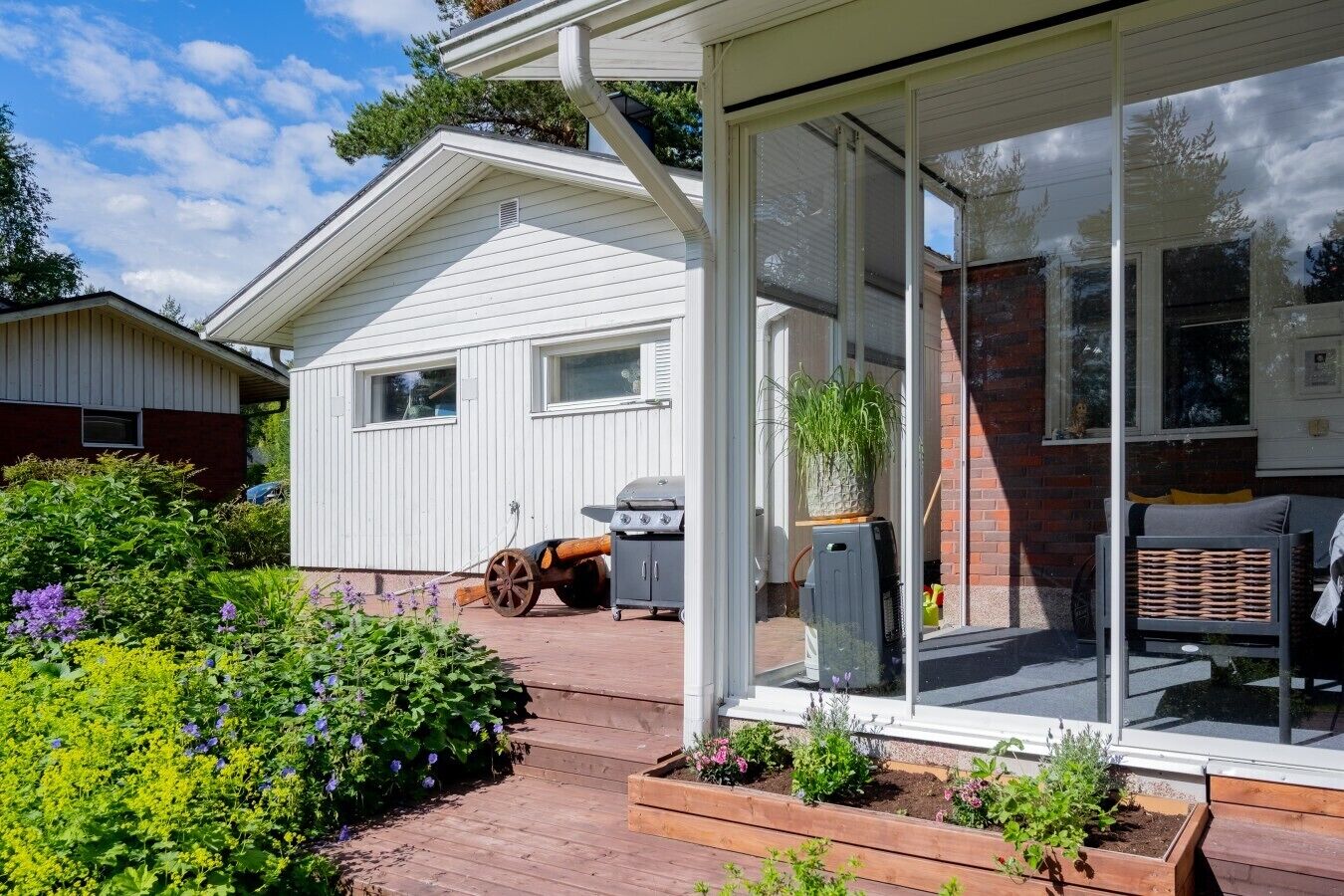 Et hus med en overdækket terrasse, grill og en have med forskellige planter og blomster.