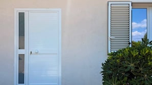 A building with a closed white door, a window with closed blinds, and a small bush in front.