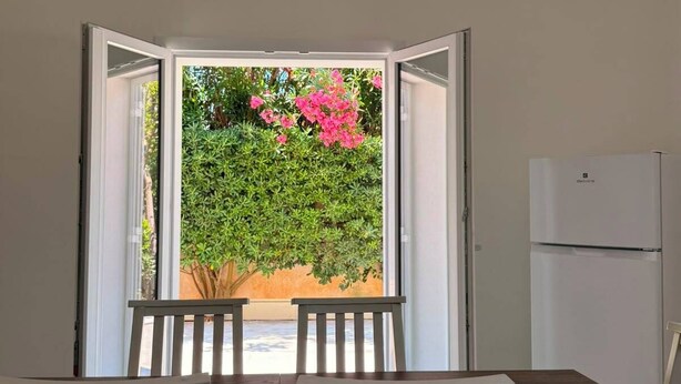 A room with a table and chairs, a window with an open door, and a refrigerator.